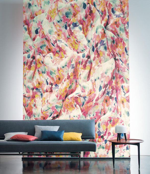 Bunt gemusterte Wand mit abstrakten, fließenden Formen in Rosa, Gelb, Blau und Weiß hinter einem dunkelblauen Sofa mit bunten Kissen und einem kleinen runden Tisch mit weißer Lampe