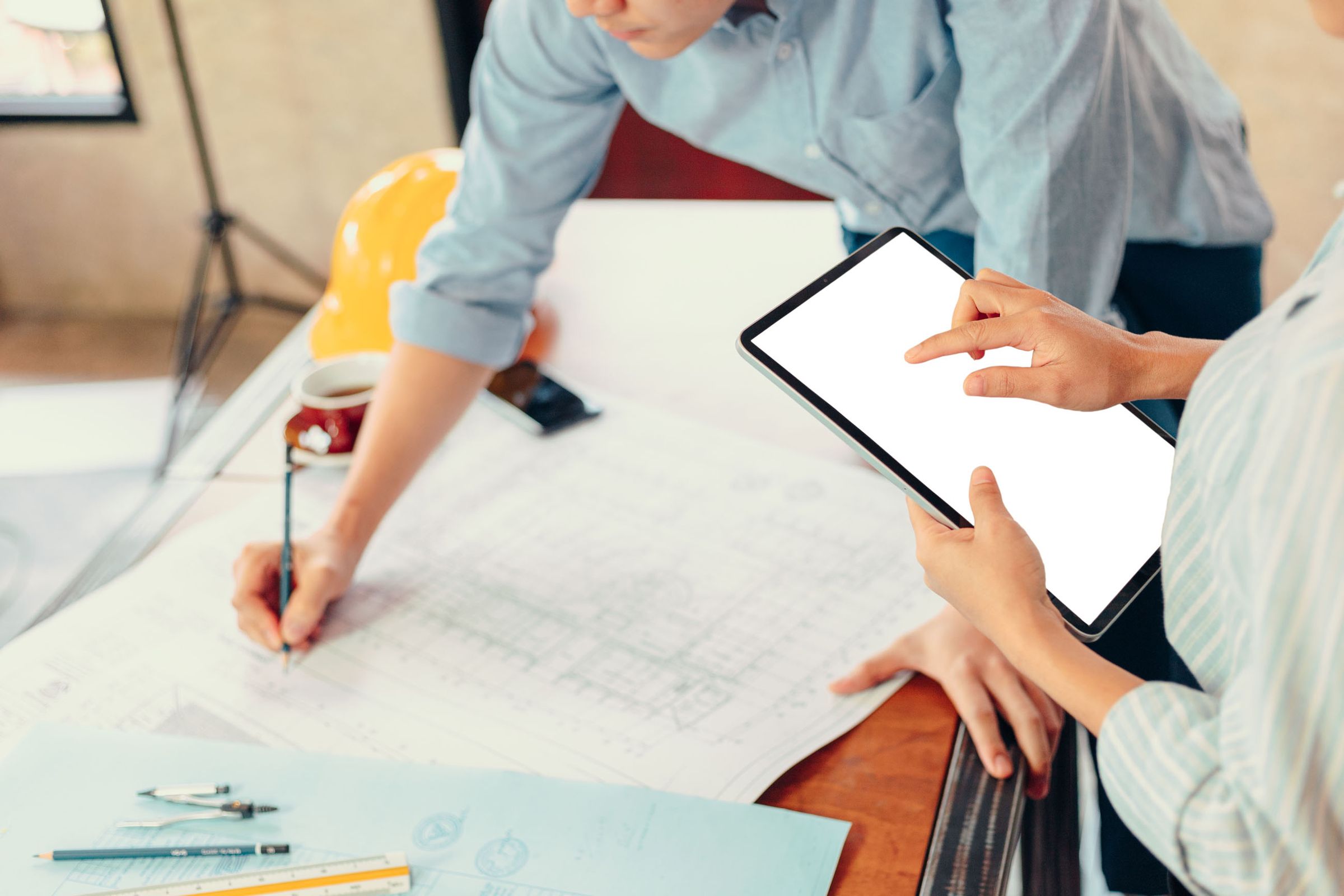 Zwei Personen arbeiten an einem Tisch mit Bauplänen, eine hält ein Tablet mit weißem Bildschirm, die andere schreibt auf den Plänen